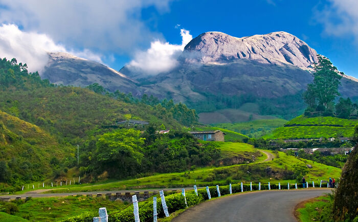 Scenic remote location with winding road, lush green hills, and a large mountain under a partly cloudy sky.