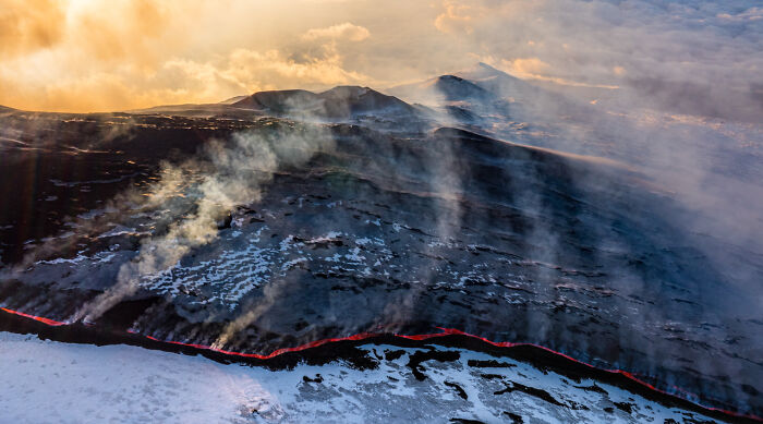 Aerial view of glowing lava flow and smoke over snow-covered volcanic landscape, showcasing award-winning nature photography.