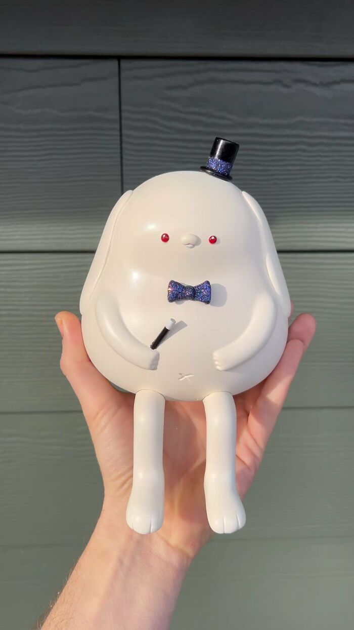 Hand holding a quirky sculpture of a white round character with red eyes, top hat, and bow tie bursting with personality.