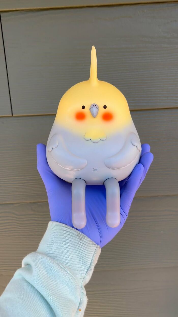 Hand holding a quirky sculpture of a chubby yellow and gray bird with long legs, bursting with personality.