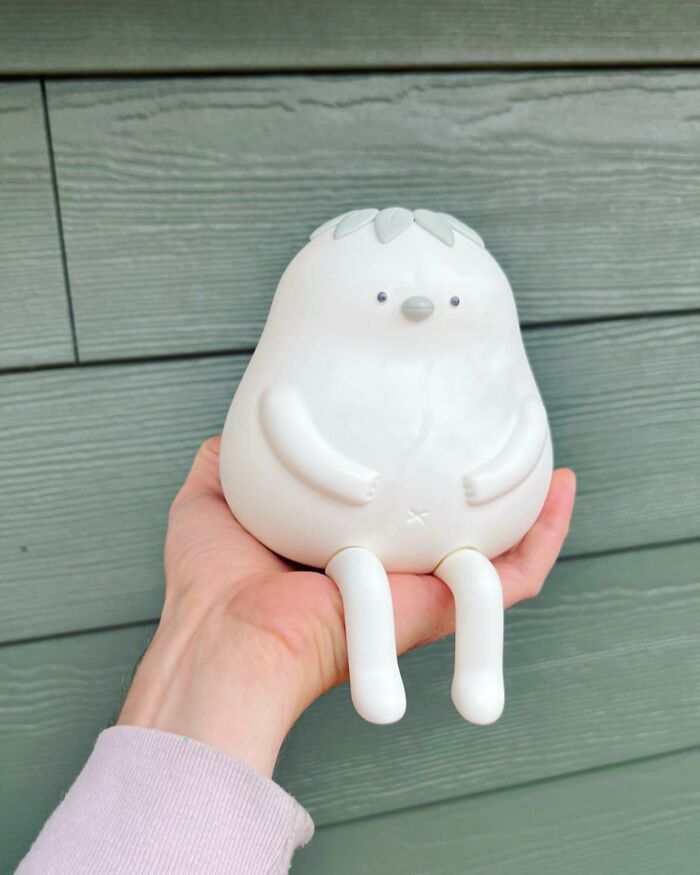 Hand holding a quirky white sculpture with personality featuring simple facial details and small legs against wooden background