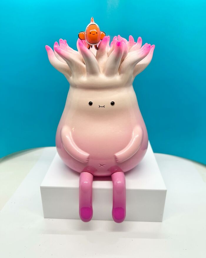 Pink quirky sculpture with a small fish on top, sitting with legs extended against a blue background, bursting with personality.