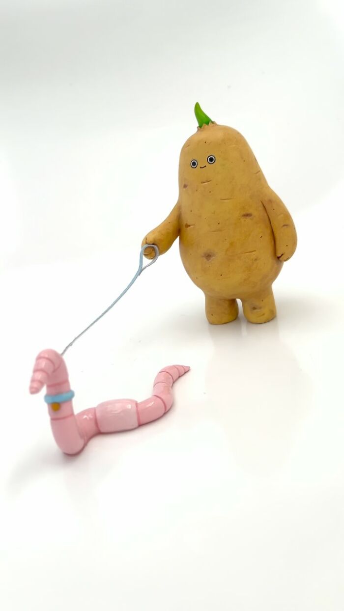 Quirky sculpture of a potato character walking a smiling pink segmented worm on a leash in a white minimal background.