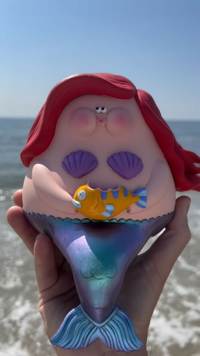Hand holding a quirky mermaid sculpture with red hair, purple shell top, and a small orange fish at the beach background.