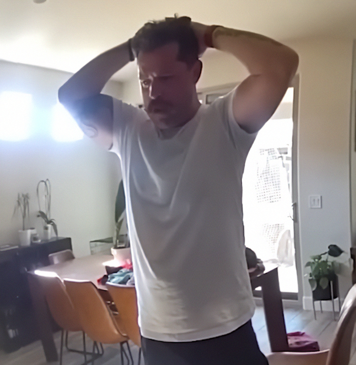 Man in white t-shirt holding his head in a living room, related to dad who fatally left 2YO daughter in car case. Man in white t-shirt holding his head in a living room, related to dad who fatally left 2YO daughter in car case.