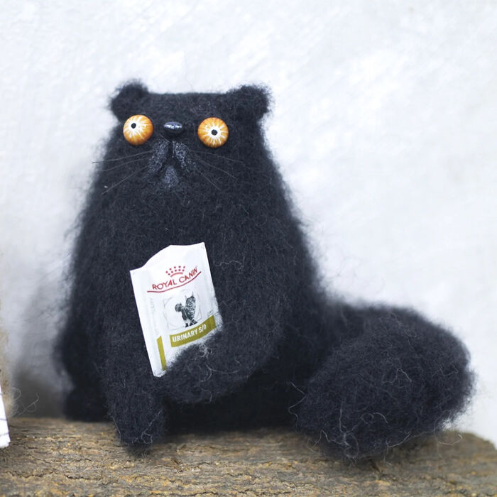 Handmade strange creature toy resembling a black fluffy cat with large eyes holding a tiny packet.