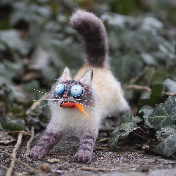 Handmade creature toy resembling a quirky striped cat with big blue eyes and an orange fish in its mouth outdoors.