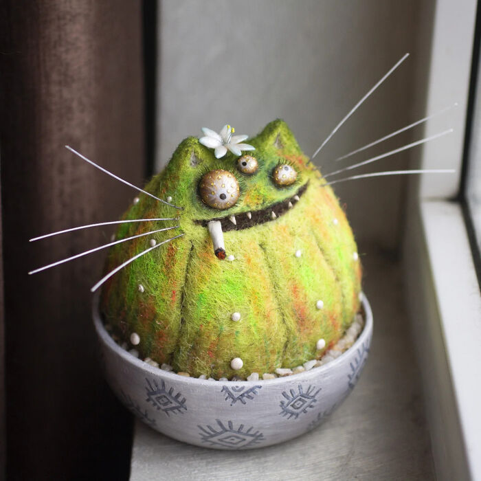 Adorably strange handmade creature toy with green fuzzy texture, big eyes, whiskers, and a small white flower on its head.