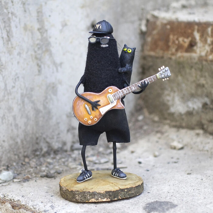 Handmade creature toy wearing a cap and glasses, holding a guitar with a small black cat on its shoulder.