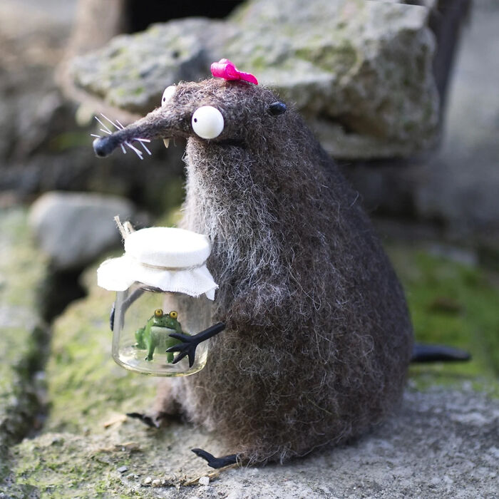 Adorably strange handmade creature with large eyes and pink bow, holding a jar with a tiny frog toy outdoors.