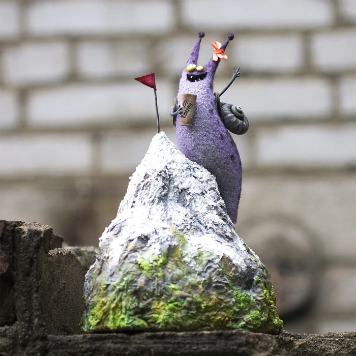Purple handmade creature toy holding a coffee cup, standing on a snowy mountain with a red flag and flower accessory.