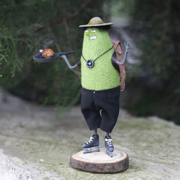 Handmade creature toy with green body, black pants, roller skates, hat, and backpack, showcasing adorably strange design.