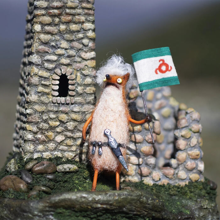 Handmade creature toy with fluffy texture holding a flag, standing by stone ruins in an outdoor setting.
