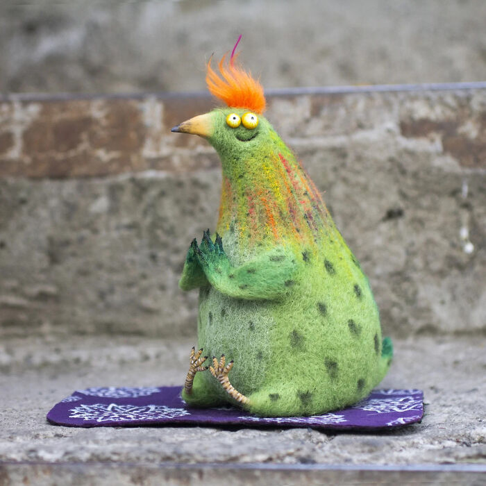 Handmade creature toy with green body, orange tuft, and spotted pattern sitting on a purple cloth outdoors.