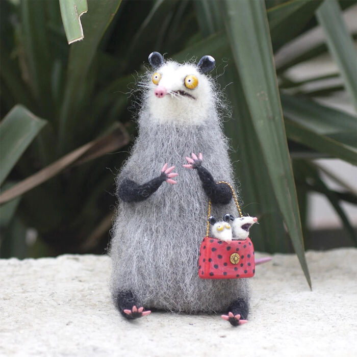 Handmade creature toy with gray fur and yellow eyes holding a red bag with two smaller handmade creatures inside.