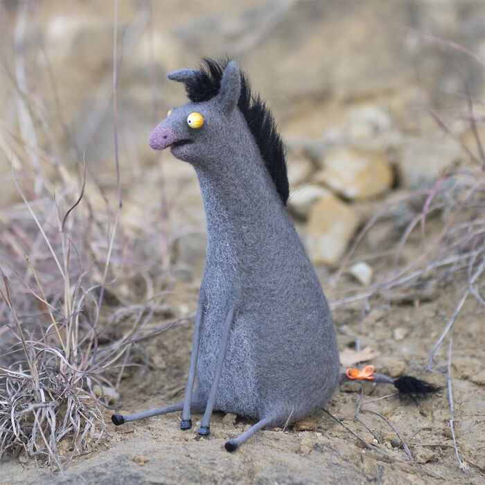 Handmade creature toy with a quirky design, featuring gray felt, yellow eyes, and black hair sitting outdoors on dirt.