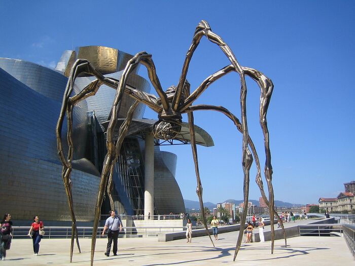 Giant spider sculpture outside modern architectural museum with visitors walking nearby on a clear sunny day.