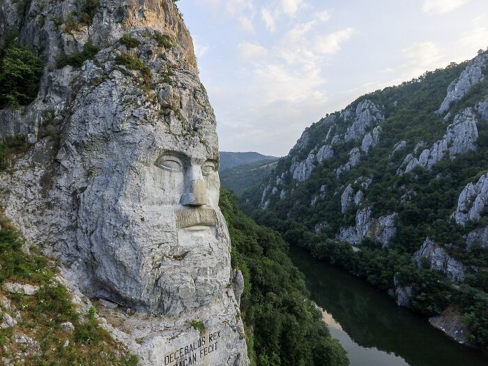 Massive rock sculpture of a bearded face carved into a cliff overlooking a river and lush green mountainous landscape.
