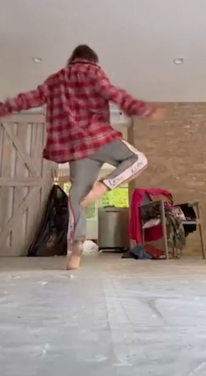 Taryn Manning dancing barefoot indoors wearing a red plaid shirt and gray sweatpants in a casual home setting.