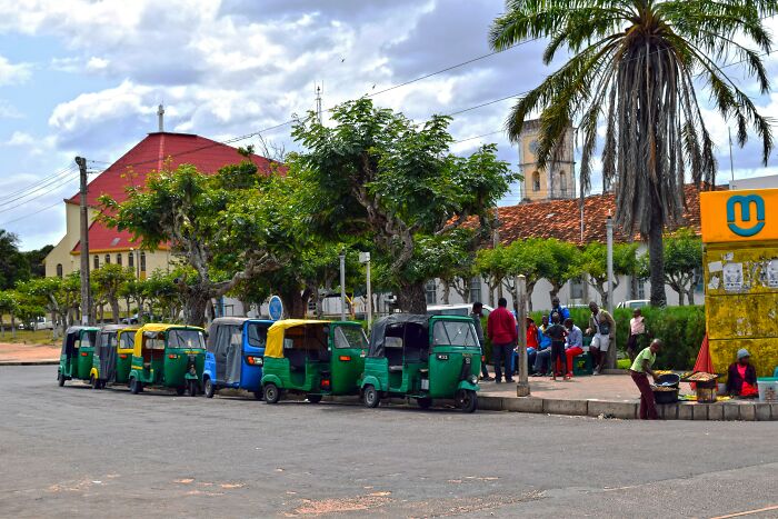 A row of colorful tuk-tuks parked on a street with people and trees in the poorest country facing hardship.