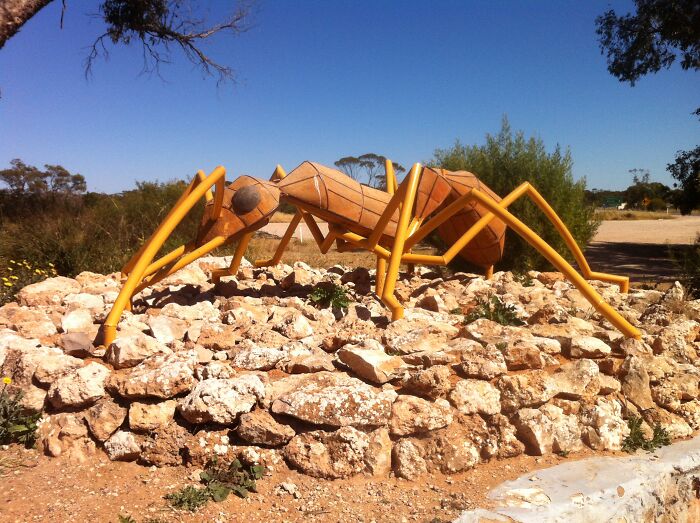 Large spectacular ant sculpture made of wood and metal displayed outdoors on rocky terrain under clear blue sky.