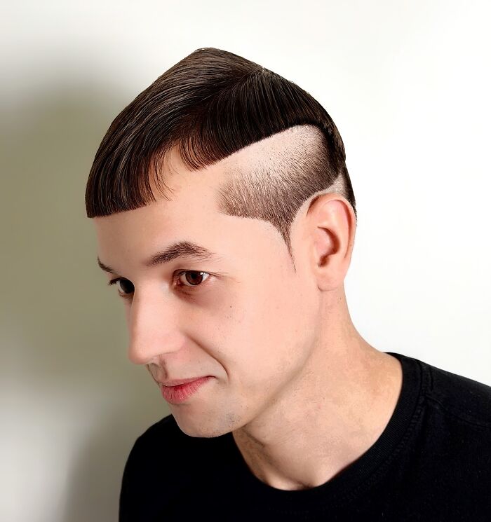 Man with a bold barber haircut featuring a sharp geometric design and textured fringe, wearing a black shirt against a white background.