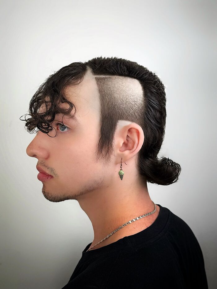 Young man with bold barber haircut featuring shaved sides, long curly fringe, and a small tied ponytail at the back.