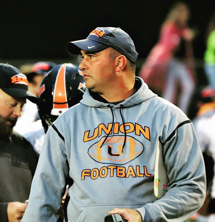 Football coach in Union Football hoodie and cap, holding playbook during a game, highlighting missing coach case mystery. Football coach in Union Football hoodie and cap, holding playbook during a game, highlighting missing coach case mystery.
