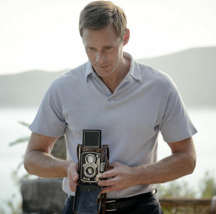 Alexander Skarsg&aring;rd holding a vintage camera, showcasing a unique detail in his otherwise conventional look outdoors.