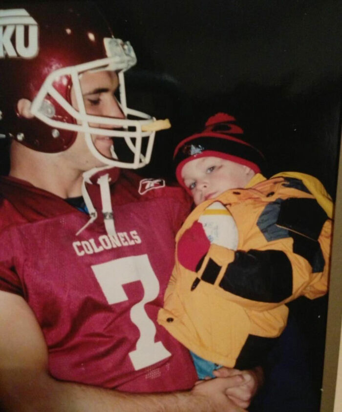 Football coach in maroon Colonels jersey holding a young child in a yellow jacket and winter hat. Football coach in maroon Colonels jersey holding a young child in a yellow jacket and winter hat.