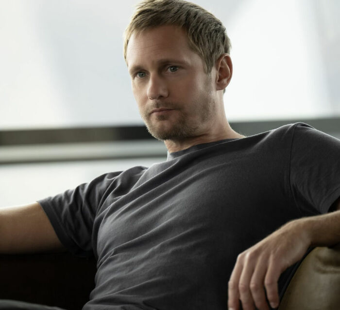 Alexander Skarsg&aring;rd in a casual dark t-shirt, sitting thoughtfully with a relaxed posture in a softly lit room.