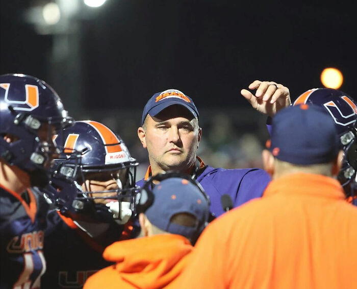 Football coach in navy and orange team gear speaking to players during a nighttime practice session. Football coach in navy and orange team gear speaking to players during a nighttime practice session.