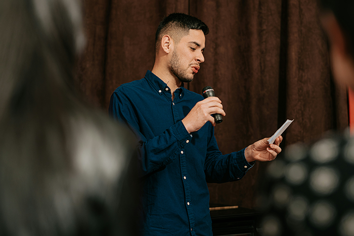 Man holding a microphone and note speaking in front of a small audience, expressing caretaker frustration and guilt trips.