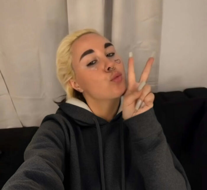 Amanda Bynes posing indoors wearing a dark hoodie, making a peace sign with her fingers and a playful expression.