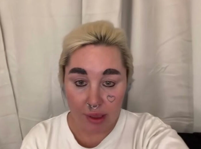 Amanda Bynes speaking in a video with a heart tattoo on her cheek and bleached hair against a white curtain background