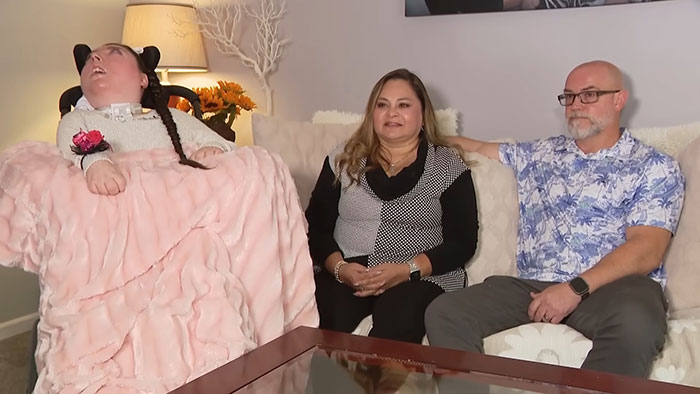Woman born without most of her brain sits with family on couch, sparking discussion about her life and condition.