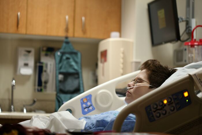 Patient lying in hospital bed with medical equipment nearby, captured in a surveillance camera image.