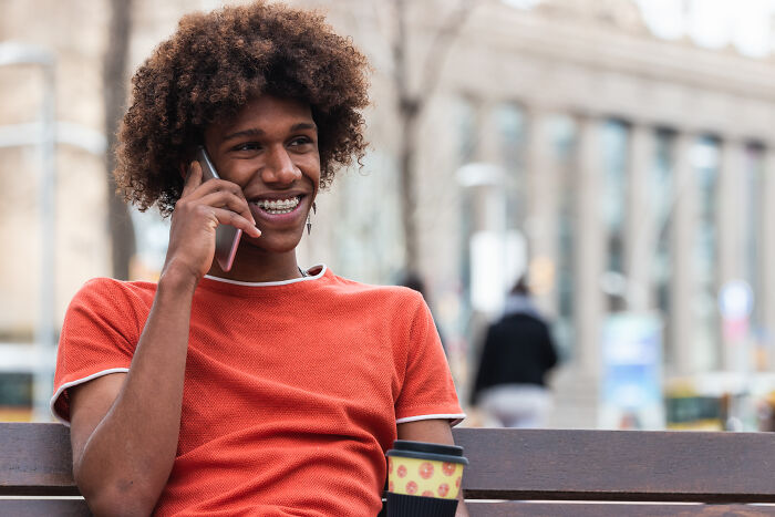 Young man smiling while talking on phone, holding coffee cup, enjoying a relaxed moment in the US outdoors.