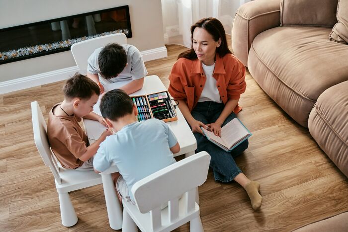 Woman reading a book to children at a small table, as they engage in childhood habits and creative activities.