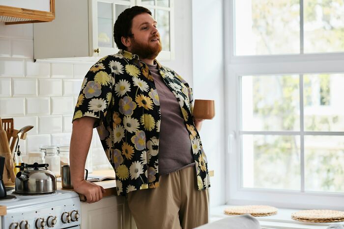 Man in floral shirt leaning on kitchen counter with mug, capturing moments when folks accidentally hurt loved ones with humor.