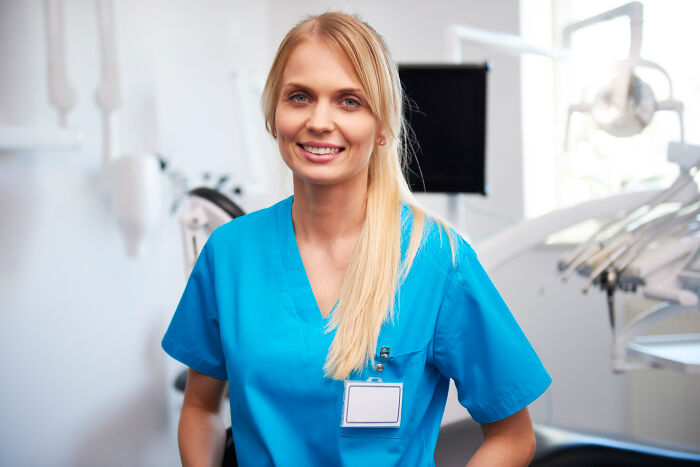 Smiling female nurse in blue scrubs standing in a bright medical room related to anesthesia patient care.