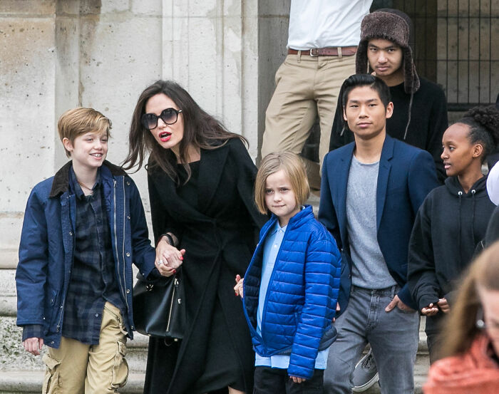 Knox Jolie-Pitt wearing a blue puffer jacket, standing with family and friends in a casual outdoor setting.
