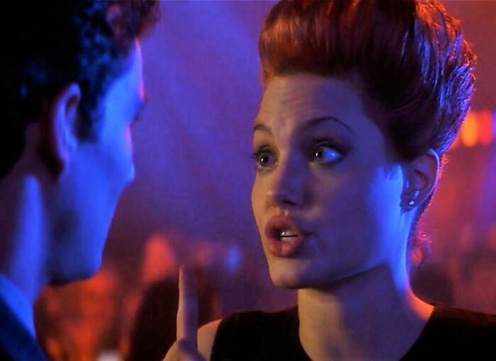 A close-up of Angelina Jolie speaking intently in a dimly lit setting with vibrant blue and orange lighting.