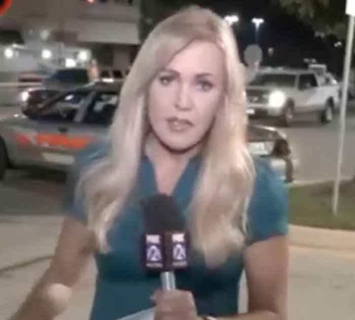 Former news anchor reporting live outdoors at night holding a microphone with police cars in the background.