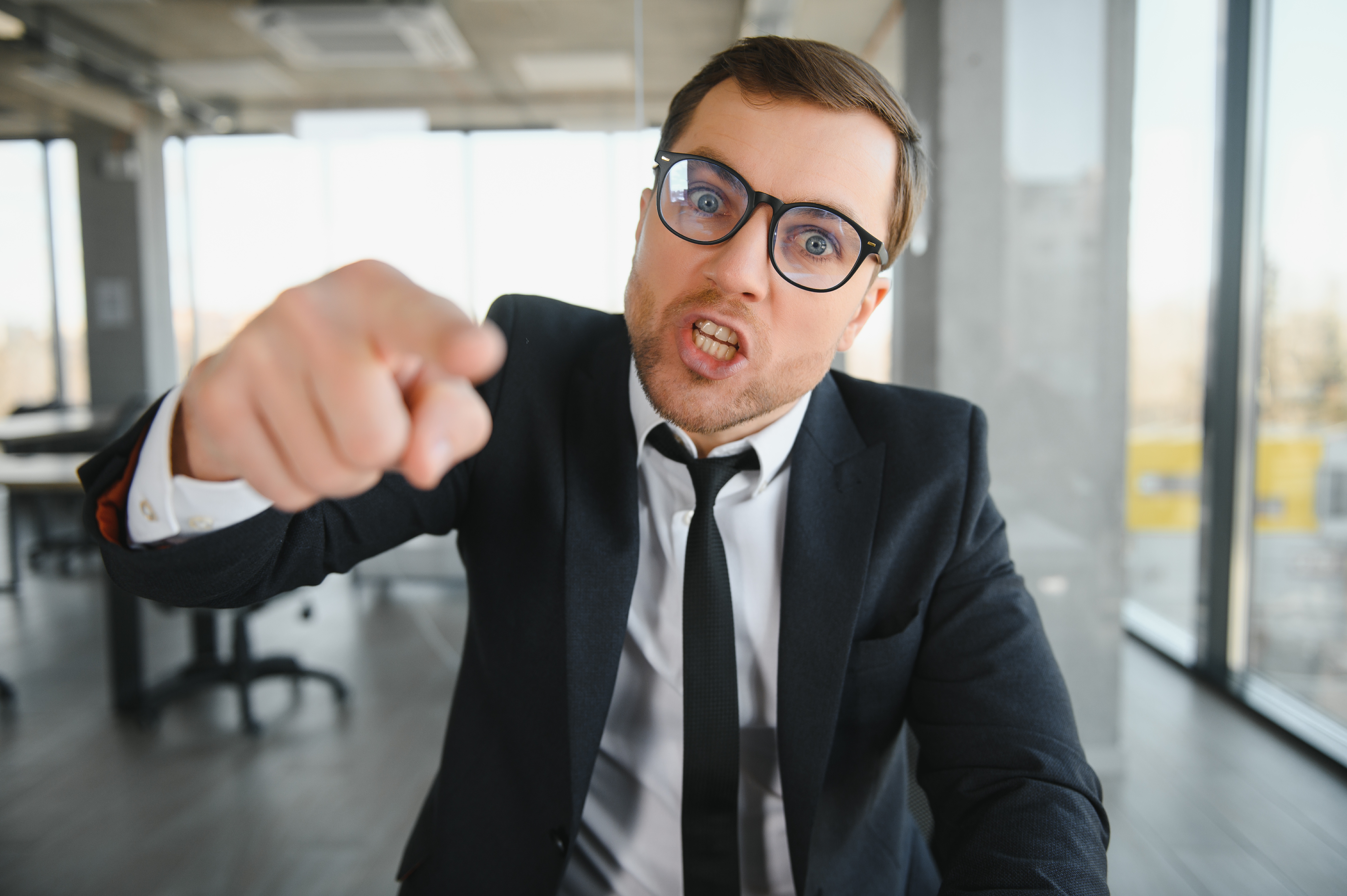 Angry lawyer in glasses and black suit pointing aggressively during a corporate meeting in a modern office.