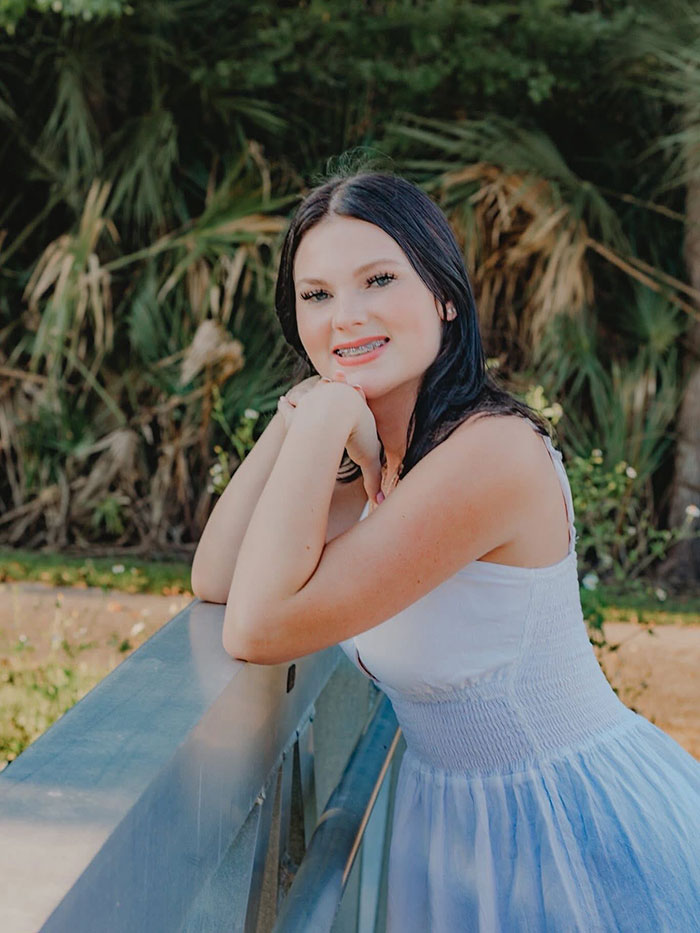 Teen stepsibling under investigation after body found stuffed under bed on Carnival Cruise, young woman posing outdoors in white dress.