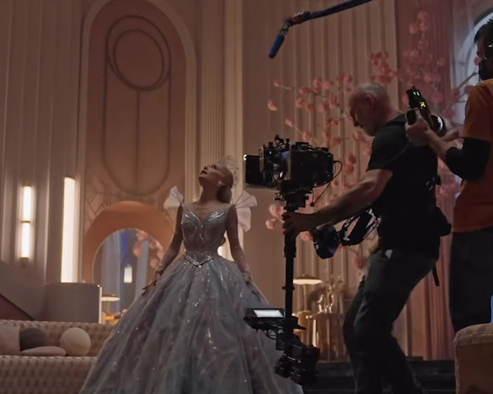 Ariana Grande filming a scene in an elegant gown with a camera crew capturing her cryptic last hurrah announcement.
