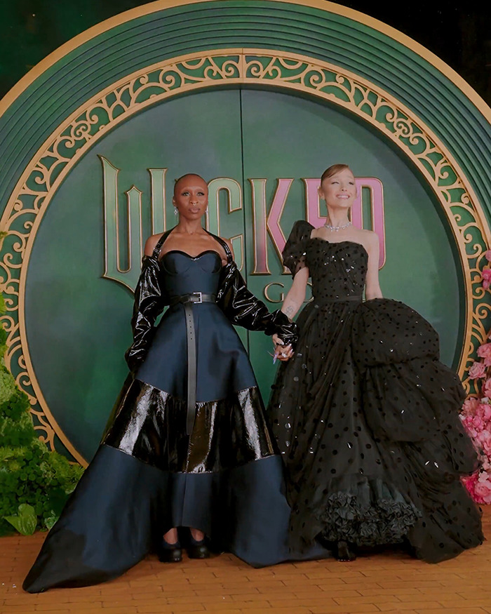Two women in elegant black gowns posing in front of a large Wicked musical logo backdrop at an event appearance.
