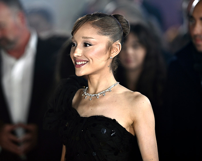 Ariana Grande smiling at an event wearing a black dress and diamond necklace, fans concerned about Wicked role impact.
