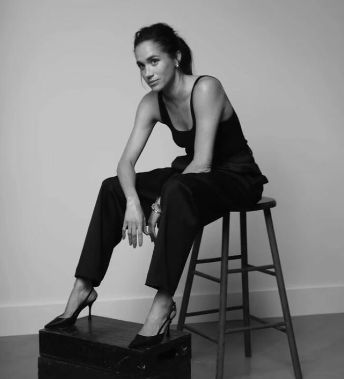 Meghan Markle in a makeup-free black outfit, sitting on a stool with a pensive expression in a minimalist setting.